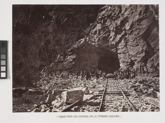 A black-and-white photograph of railroad tracks leading into a rocky tunnel, with workers gathered at the  tunnel entry.