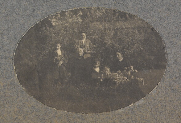 An oval-shaped black-and-white photograph of a group of light-skinned women and children holding flower bouquets in a natural setting.