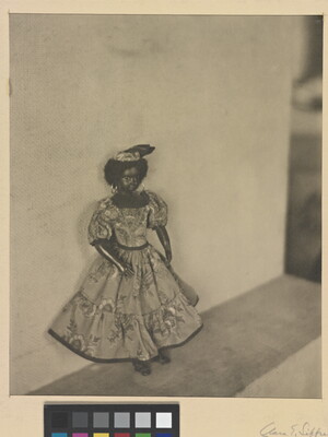 A black-and-white photograph of a dark-colored doll with curly hear wearing a patterned dress and hat posed against a wall.