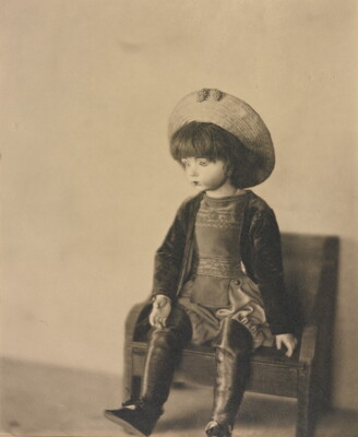 A sepia-toned photograph of a light-colored doll wearing a hat and boots sitting on a small bench.