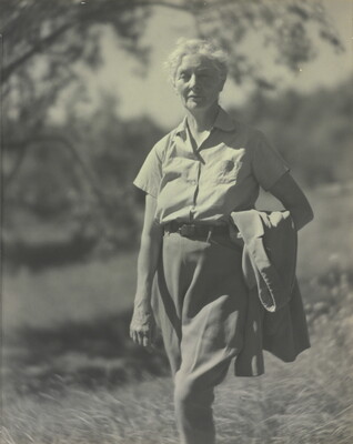 A black-and-white photograph of an older White woman with short, white hair standing outside holding a jacket in the crook of her arm.