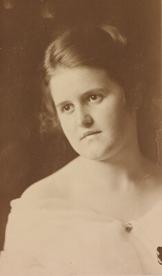 A sepia-toned portrait photograph of a White woman, her head slightly tilted and hair pulled up, wearing an off-the-shoulder dress.