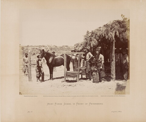 A black-and-white photograph of several people and one horse standing in a dirt yard outside of a hut with leafy branches as a roof.