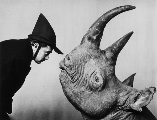 A black-and-white photograph of a White man wearing a Peter Pan-style hat facing a rhinoceros; both human and animal in profile.