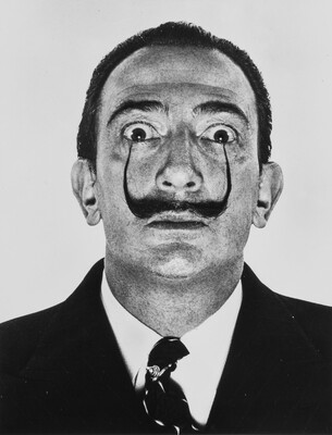 A black-and-white portrait photograph of a White man with a pencil mustache, the tips curved up all the way to his wide eyes.