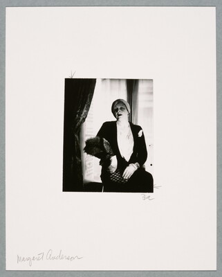 A black-and-white photograph of a White woman, hair wrapped in a turban, seated near a window wearing a black jacket, white gloves, and a fur stole.