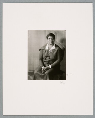 A black-and-white photograph of a seated Black woman, hair wrapped in a turban, wearing a dark suit-dress, hands folded in her lap.