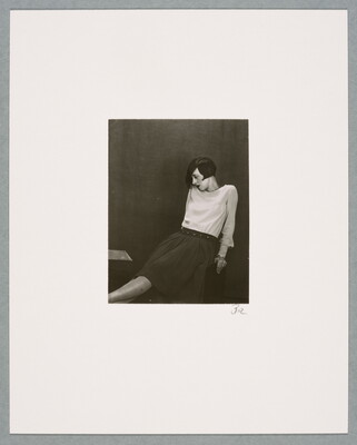 A black-and-white photograph of a seated White woman with dark pageboy hair, wearing a white blouse and dark skirt, leaning back with her legs crossed.