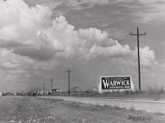 A black-and-white photograph of a rural road lined with billboards and powerlines, and a rollercoaster in the distance.