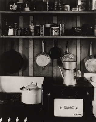 A black-and-white photograph of an old black stovetop with a white pot, a tea kettle, and shelves in the background filled with old-fashioned kitchen essentials.