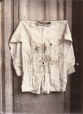 A black-and-white photograph of a shirt with bullet holes and bloodstains hanging on a door.
