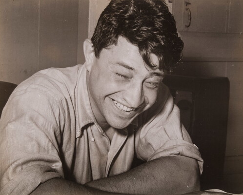 A black-and-white photograph of a seated White man laughing and leaning forward on his arms.
