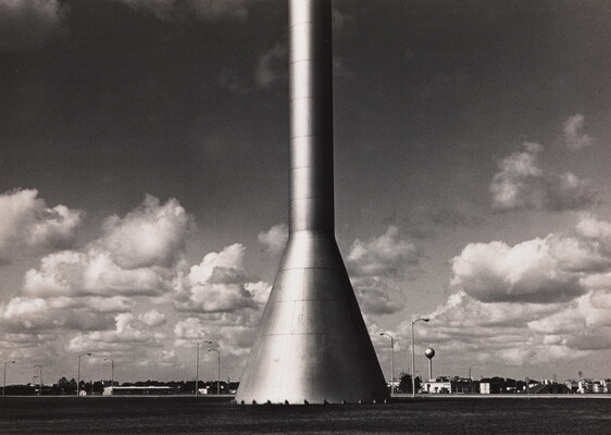 A black-and-white photograph of the bottom of a rocket-shaped tower; in the background are parking lots and streetlights.