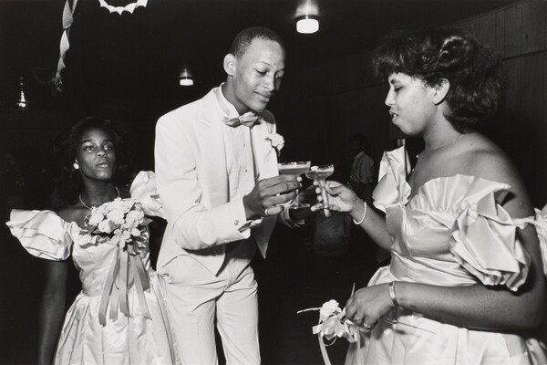 A black-and-white photograph of a Black man in a white tuxedo toasting with a Black woman in a formal dress as another woman watches.