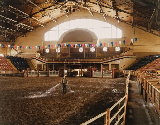 A color photograph of a man standing in an empty rodeo arena watering the dirt.