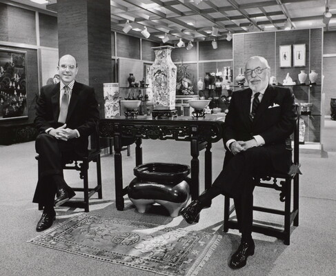 A black-and-white photograph of two smiling White men in suits sitting on either side of a table displaying Asian decor.