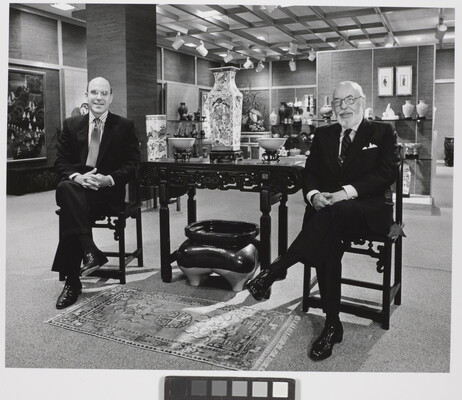 A black-and-white photograph of two smiling White men in suits sitting on either side of a table displaying Asian decor.
