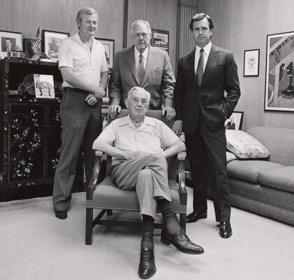 A black-and-white photograph of four White men of varying ages, three standing and one seated in an armchair, in an office.