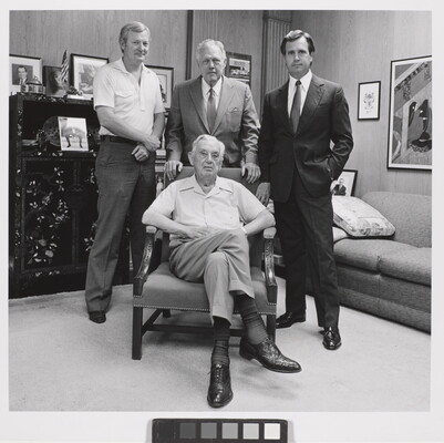 A black-and-white photograph of four White men of varying ages, three standing and one seated in an armchair, in an office.