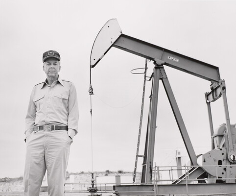 A black-and-white photograph of an older White man standing in front of a pump jack.