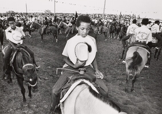 A black-and-white photograph of a large gathering of people on horses; two Black boys in the foreground holding their cowboy hats to their hearts.
