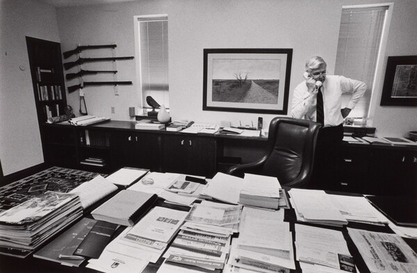 A black-and-white photograph of an older White man talking on a phone behind a large desk piled with papers.