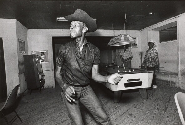 A black-and-white photograph of a Black man wearing a cowboy hat dancing in a pool hall in front of a billiards table.