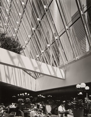 A black-and-white photograph of a cafeteria under a ceiling of windows that look up to a city skyline.