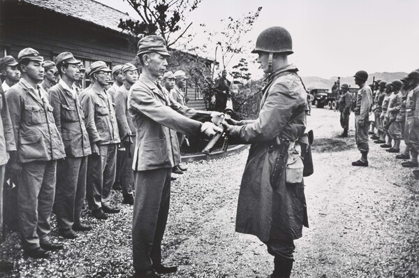 A black-and-white photograph of an Asian man handing a sword to a White man, both are in military uniforms and have groups of soldiers lined up behind them.