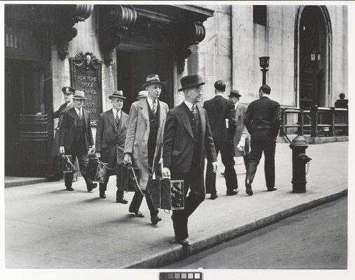 A black-and-white photograph of four White men in suits and hats exiting a city building and each holding a briefcase tethered to the other briefcases.