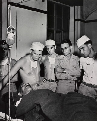 A black-and-white photograph of a man on a hospital bed hooked to an IV and under a blanket surrounded by five White men, some in military clothing, all watching him.