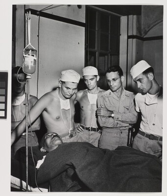 A black-and-white photograph of a man on a hospital bed hooked to an IV and under a blanket surrounded by five other White men, some in military clothing, all watching him.