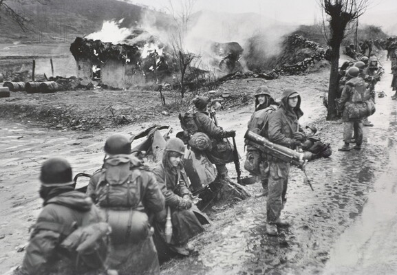 A black-and-white photograph of a line of soldiers loaded with gear on a muddy road as a building burns in the background.
