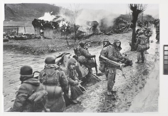 A black-and-white photograph of a line of soldiers loaded with gear on a muddy road as a building burns in the background.