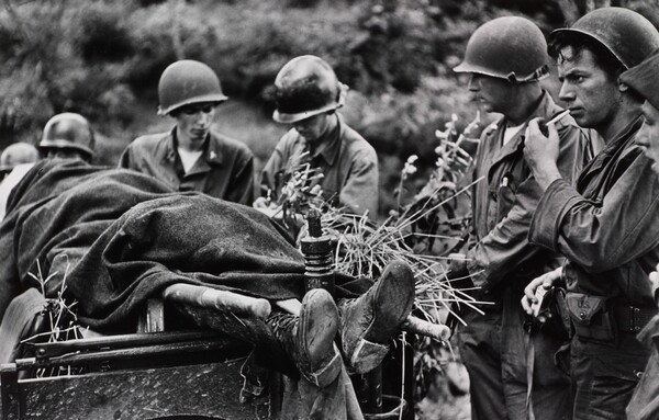 A black-and-white photograph of several helmeted White soldiers standing next a body covered in a blanket on a stretcher.
