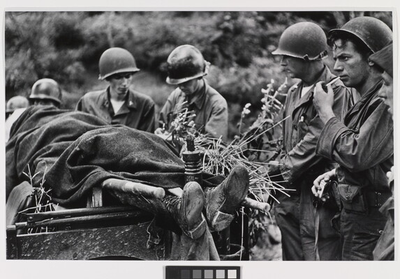 A black-and-white photograph of several helmeted White soldiers standing next a body covered in a blanket on a stretcher.