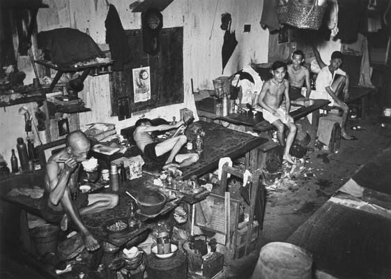 A black-and-white photograph of five emaciated Asian men wearing only shorts seated or lying on cluttered tables in a messy room.