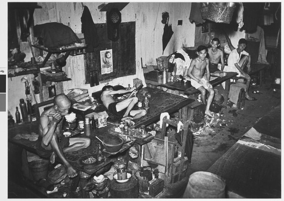 A black-and-white photograph of five emaciated Asian men wearing only shorts seated or lying on cluttered tables in a messy room.
