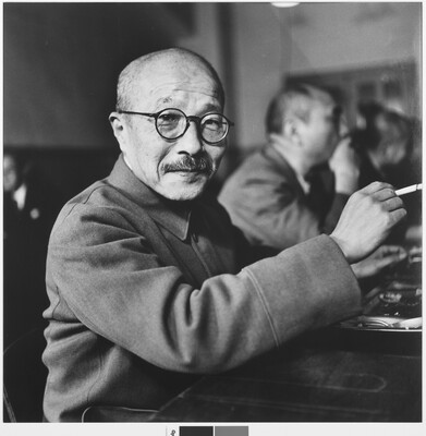 A black-and-white photograph of an Asian man with a shaved head, moustache, and round glasses holding a cigarette.