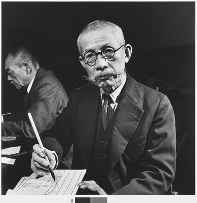 A black-and-white photograph of an Asian man with a shaved head, handlebar moustache, and round glasses writing on paper with a brush and ink.