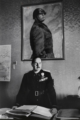 A black-and-white photograph of a White man in military regalia behind a desk stacked with papers above which hangs a portrait of a helmeted man in military dress.