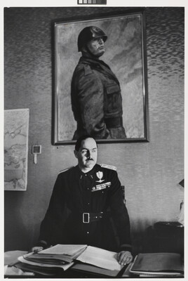 A black-and-white photograph of a White man in military regalia behind a desk stacked with papers above which hangs a portrait of a helmeted man in military dress.