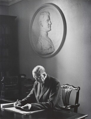 A black-and-white photograph of an older, bespectacled, White man studying papers at a desk; an oval bas-relief portrait hangs on the wall behind him.