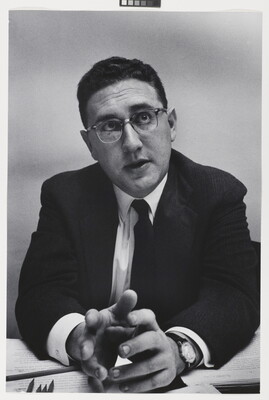 A black-and-white photograph of a White man with short, dark hair in a suit, tie, and glasses leaning forward with his elbows on a desk, gesturing with his hands.