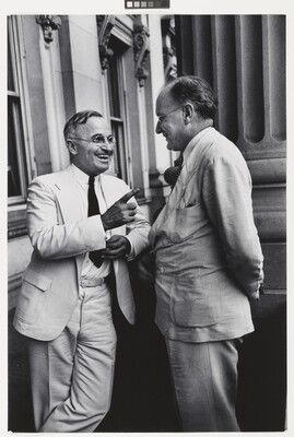 A black-and-white photograph of two White men, both in light-colored suits and glasses, talking to one another and smiling.