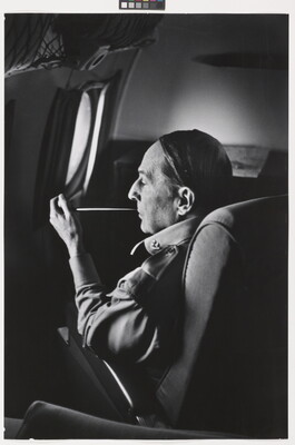 A black-and-white photograph of a White man seated on an airplane smoking a long thin pipe.