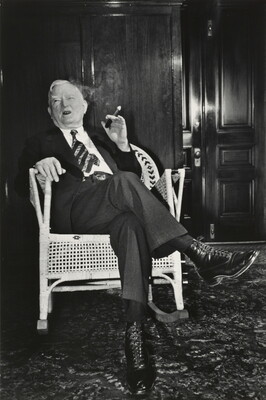 A black-and-white photograph of an older White man wearing a suit and smoking a cigar in a wicker rocking chair.