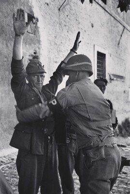 A black-and-white photograph of a White man in military uniform being frisked by a second man in military uniform.