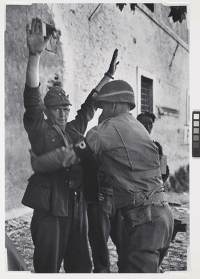 A black-and-white photograph of a White man in military uniform being frisked by a second man in military uniform.