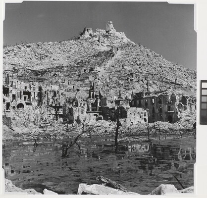 A black-and-white photograph of the ruins of a town at the edge of a body of water and a ruined building on top of a mountain in the background.